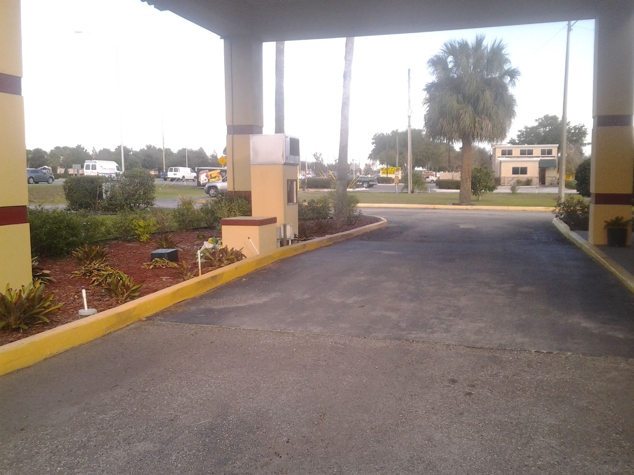 A parking area with a covered entrance, a small planter with plants, and a building in the background.