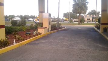 A parking area with a covered entrance, a small planter with plants, and a building in the background.