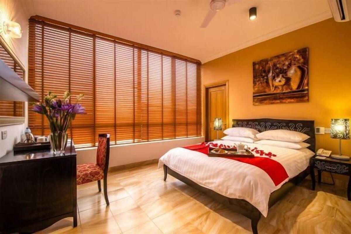 A hotel room with a bed, a desk, a chair, a window with blinds, a painting on the wall, and a nightstand with a lamp.