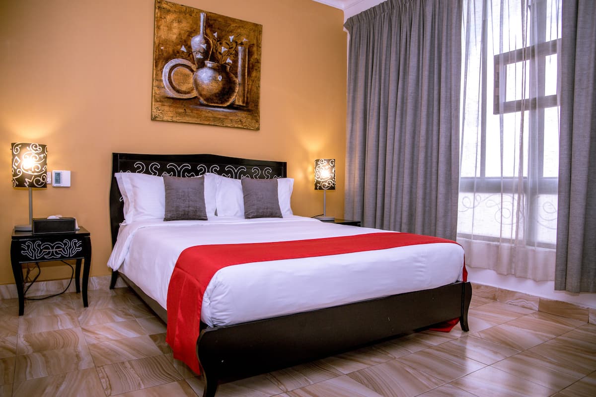 A neatly made bed with a red and white bedspread, two bedside tables with lamps, a wall-mounted artwork, and a large window with curtains.