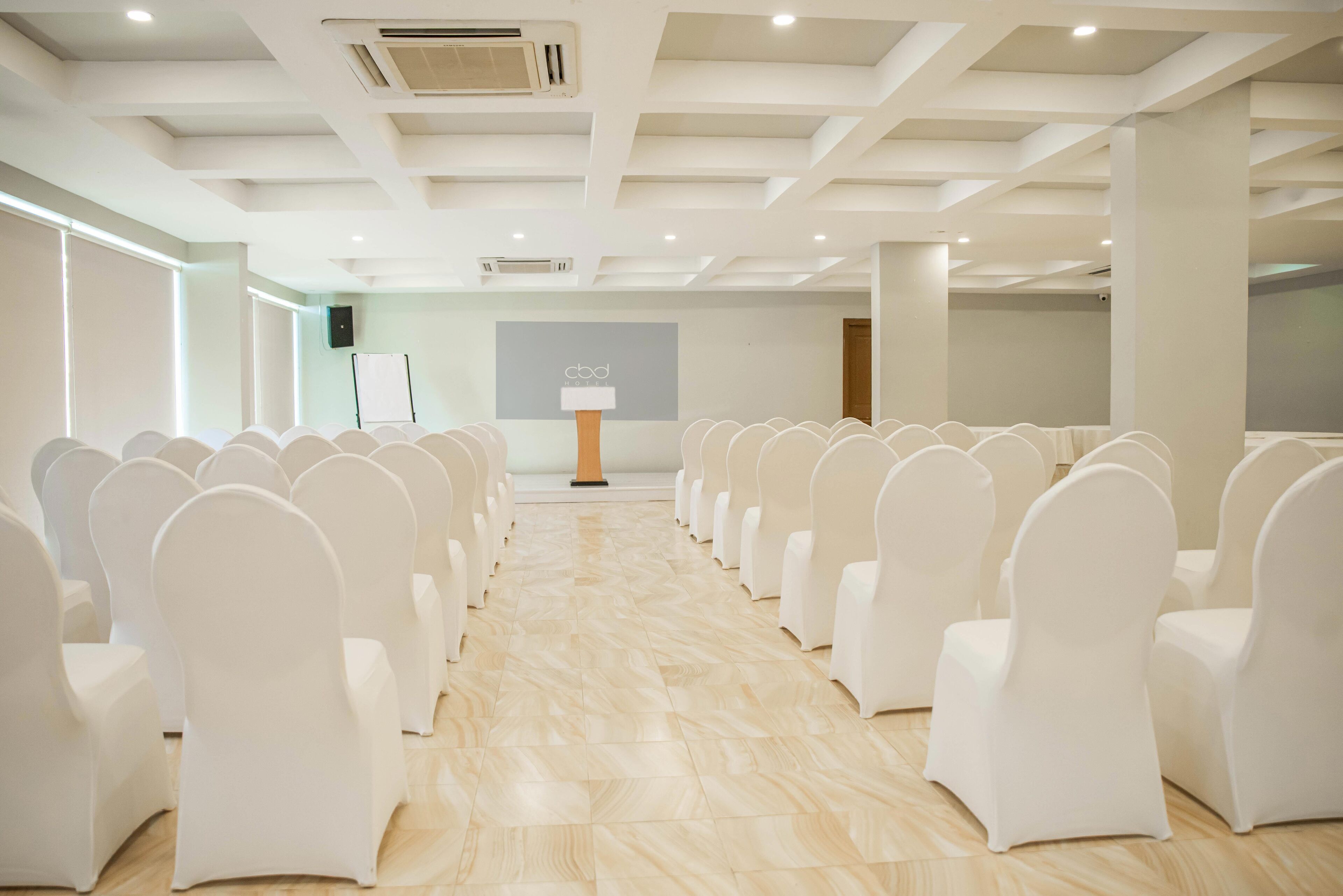 Une salle de conférence avec des rangées de chaises blanches, un écran de projection et un tableau blanc.