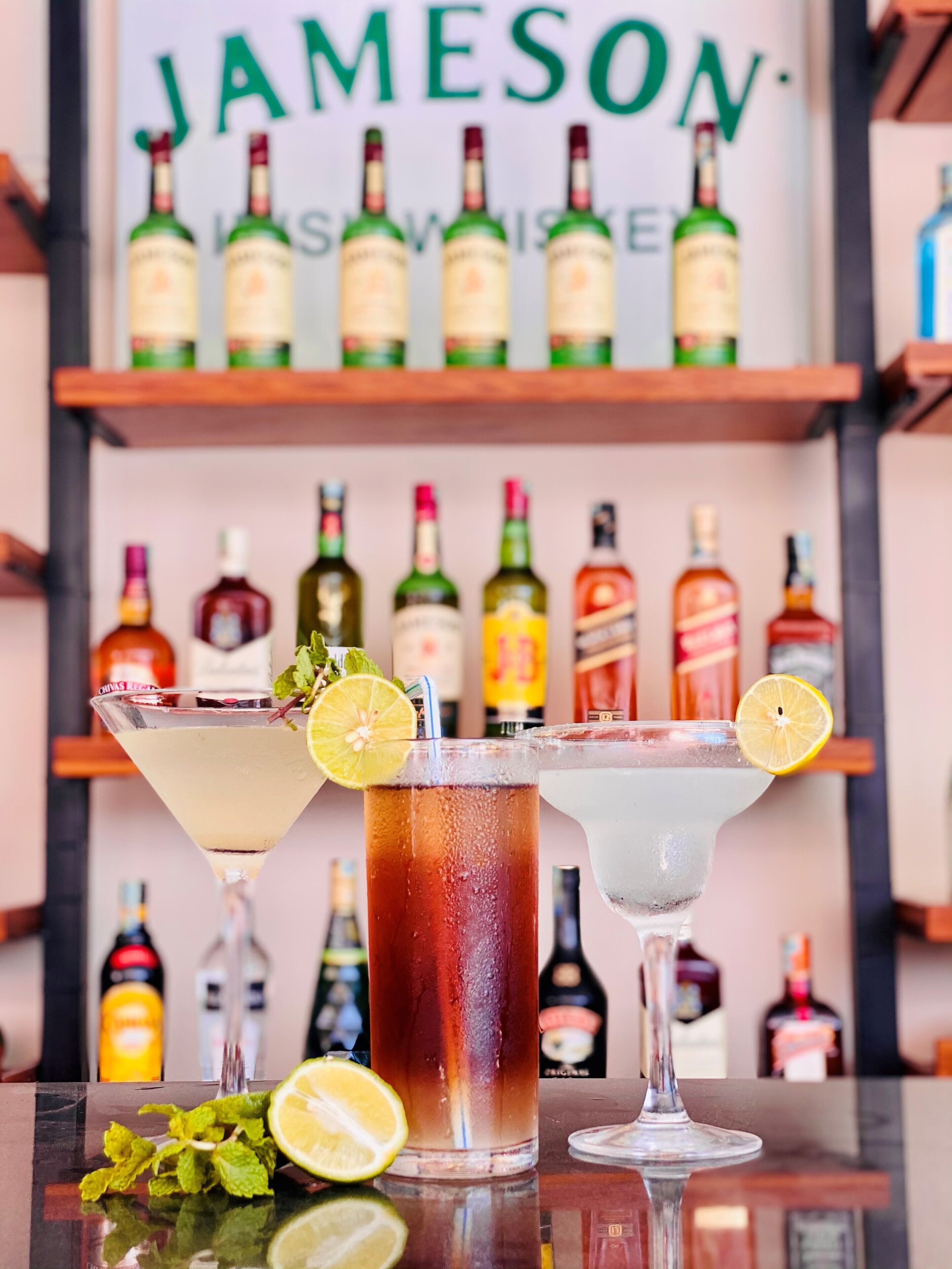 Three cocktails on a bar counter with a background of various liquor bottles.