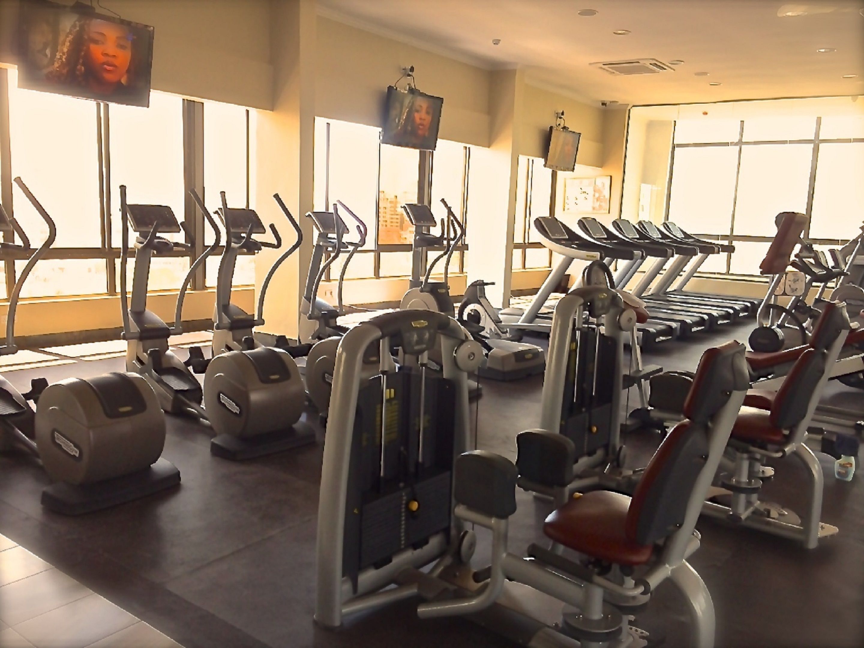 A gym with various exercise machines, large windows, and a television screen.