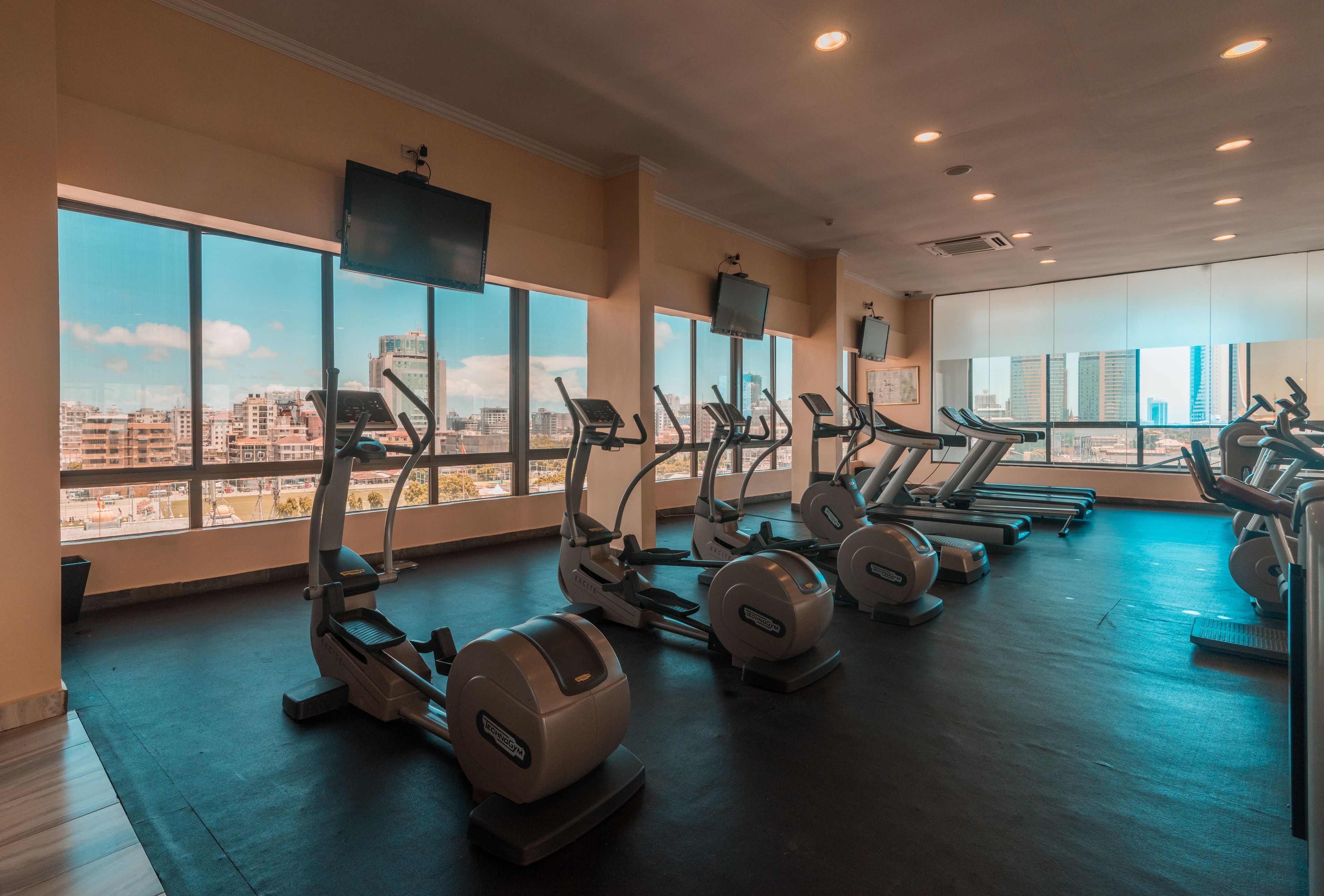A gym with multiple treadmills and elliptical machines, large windows offering a city view, and a flat-screen TV mounted on the wall.