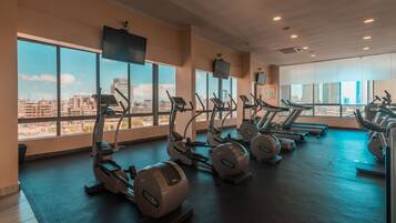 A gym with multiple treadmills and elliptical machines, large windows offering a city view, and a flat-screen TV mounted on the wall.