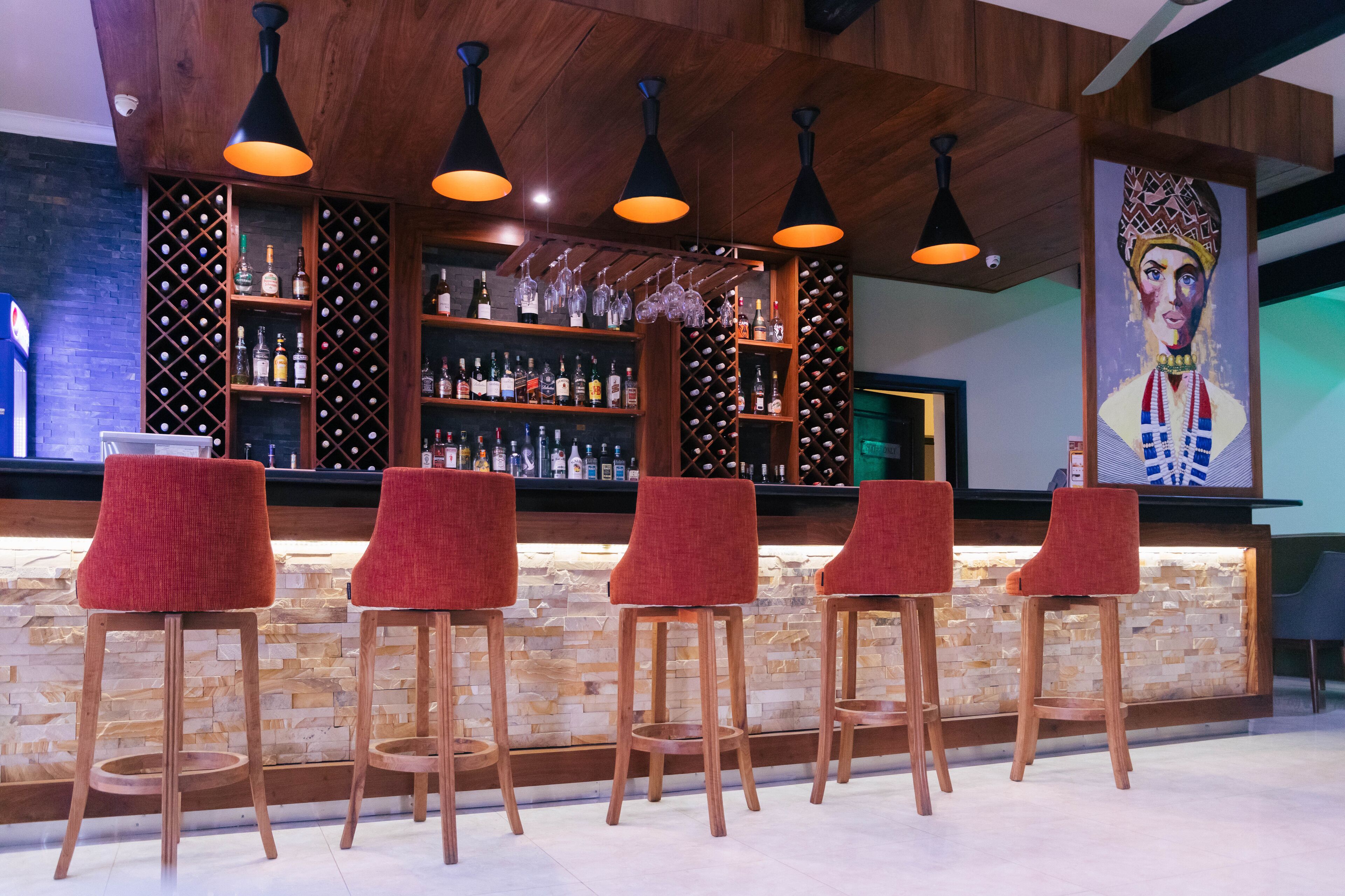 A bar area with wooden shelves, red bar stools, and hanging lights.