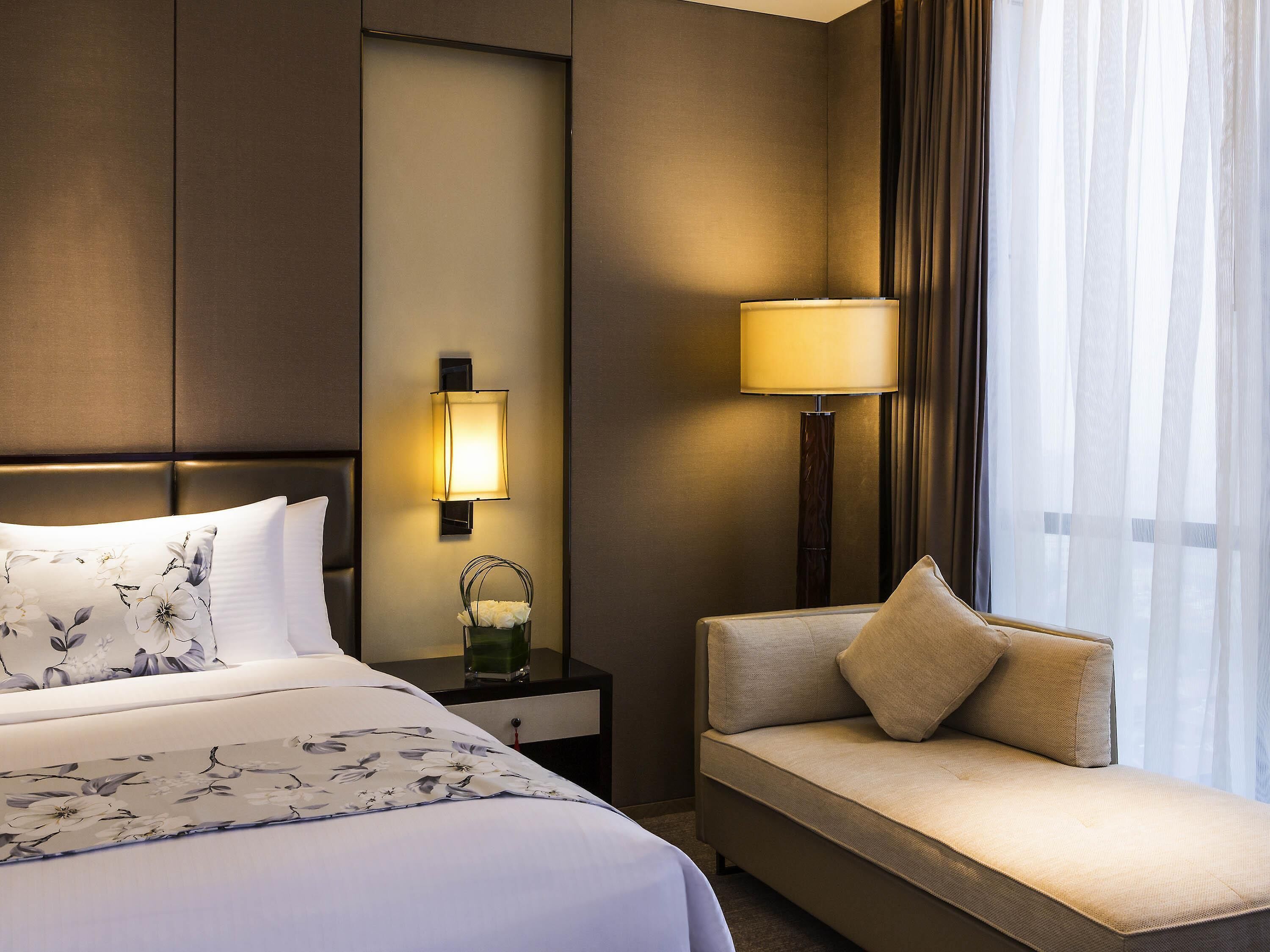 Ein Hotelzimmer mit Bett, Nachttisch, Lampe, Sofa und einem Fenster mit Vorhängen.