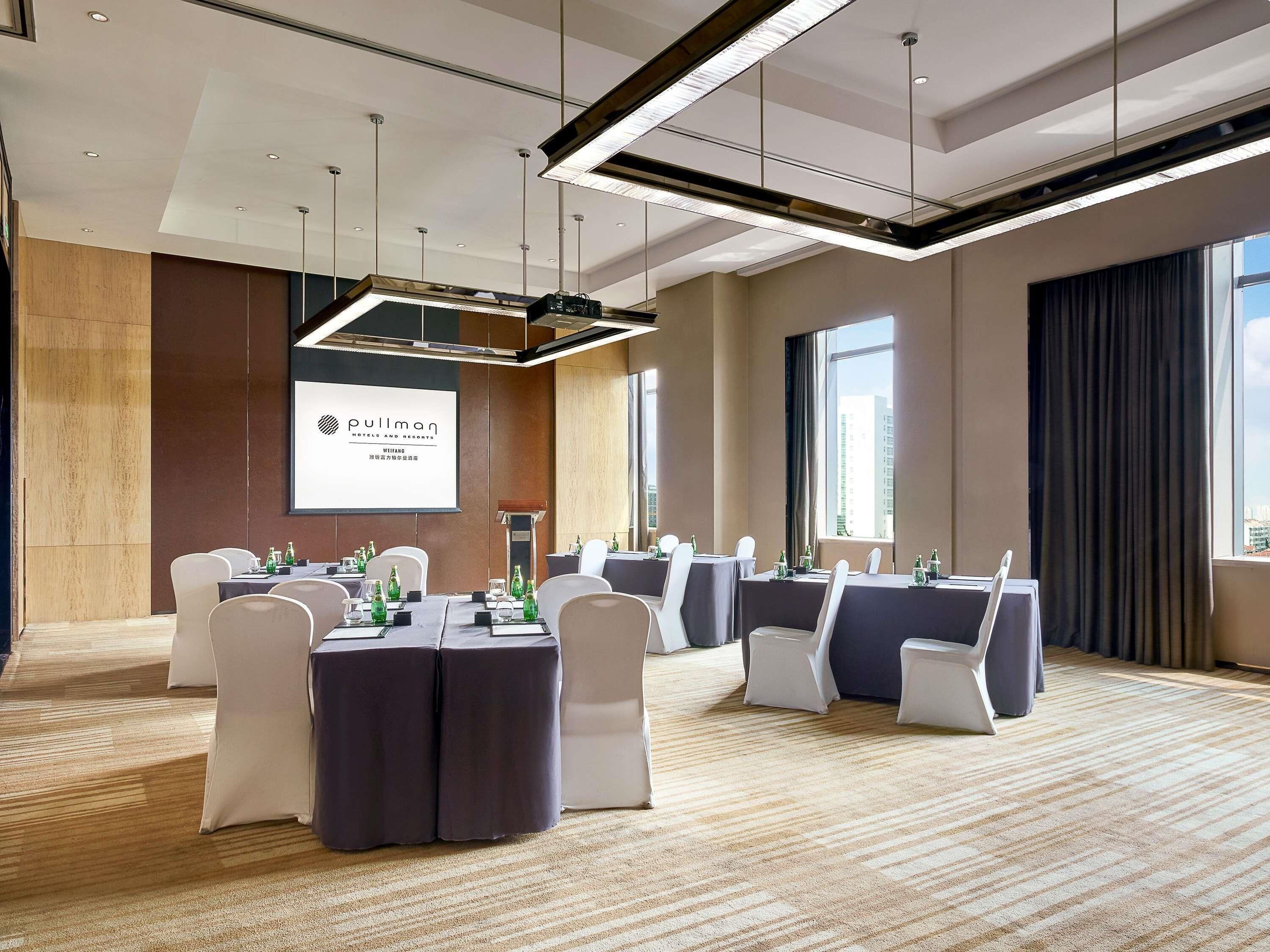Un salón de conferencias con una gran pantalla mostrando el nombre del hotel, varias mesas con sillas y piso alfombrado.