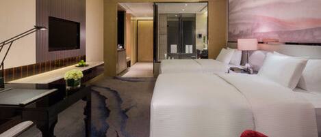 A modern hotel room with a large bed, a desk with a flat-screen TV, and a bathroom visible through a glass door.