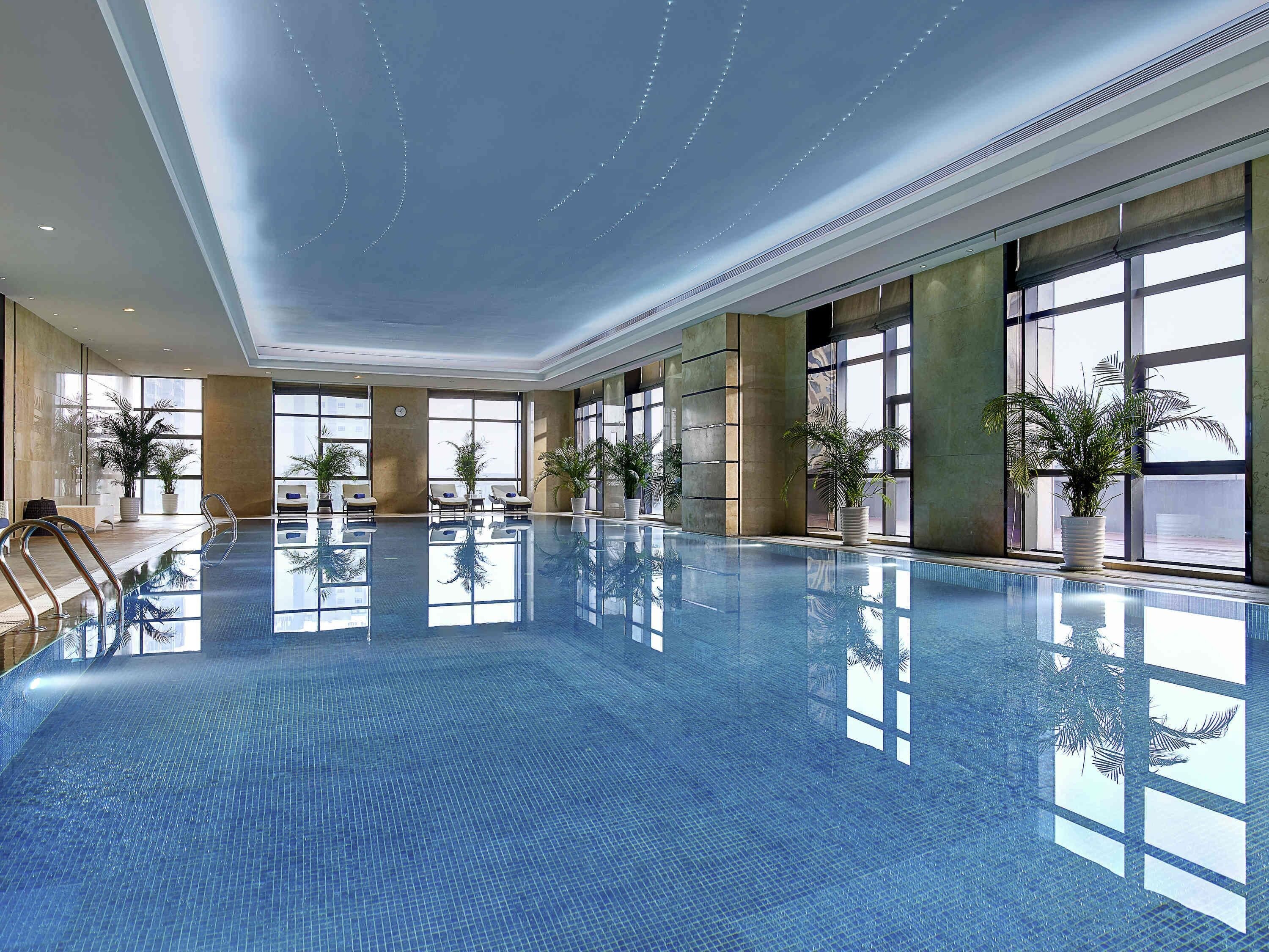A large indoor swimming pool with clear reflections, large windows, and potted plants.