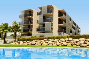 A modern multi-story building with balconies, a swimming pool, and a landscaped area with rocks and greenery. - Rentalmar Families Paradise Village (Salou)
