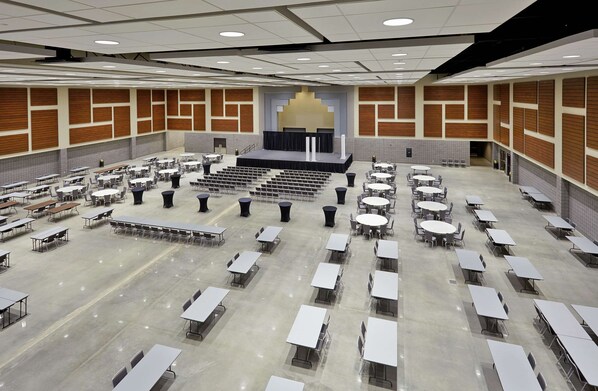 Un amplio salón de conferencias bien iluminado, con varias mesas y sillas, un escenario con un atril y suelo de baldosas.