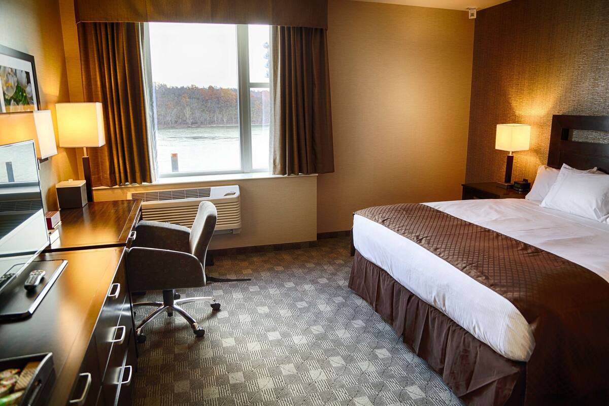 A hotel room with a bed, desk, chair, and a view of a body of water through the window.