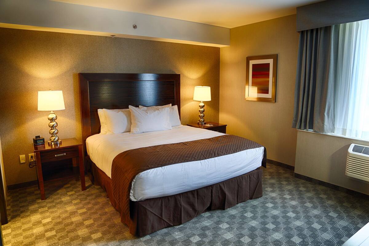 A hotel room with a large bed, two bedside tables with lamps, a window with curtains, and a wall-mounted air conditioner.