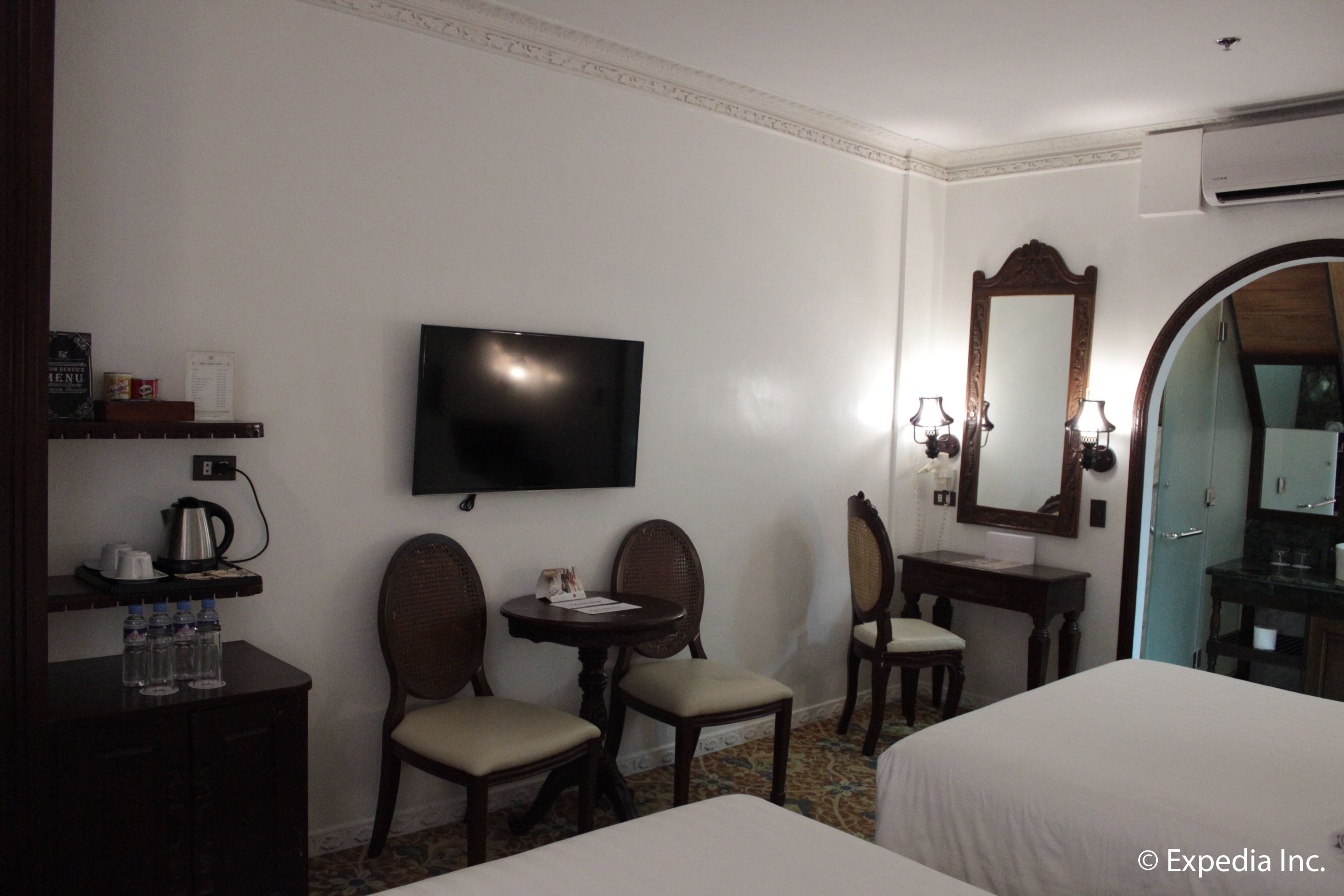 A hotel room with a bed, a TV, a desk with a telephone, two chairs, a small table, and a mirror.