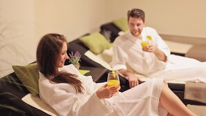 Twee personen in witte gewaden zitten op een bed met groene kussens, met glazen sinaasappelsap in hun handen.