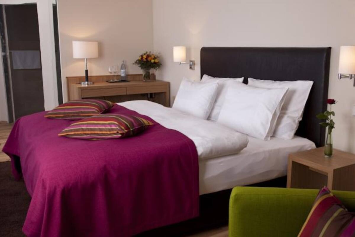 Een hotelkamer met een bed, een nachtkastje, een lamp, een vaas met bloemen en een groene fauteuil.