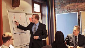 A business meeting with a man presenting a graph on a whiteboard in a room with brick walls and a window.