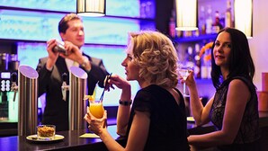 Een bar met een bartender die drankjes bereidt, een gast die een cocktail vasthoudt, en een andere gast die toekijkt.