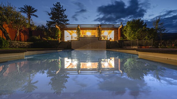 A swimming pool with a clear reflection, a covered area with seating, and surrounding greenery. - Casa Arrigo (Linguaglossa)