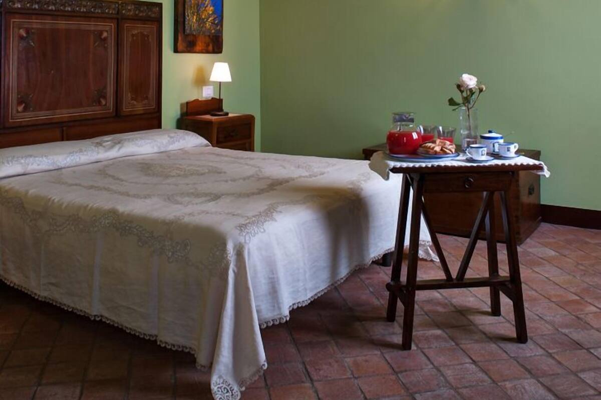 A bedroom with a wooden ceiling, a bed with a white bedspread, a nightstand with a lamp, a small table with a vase and a teapot, and a painting on the wall.
