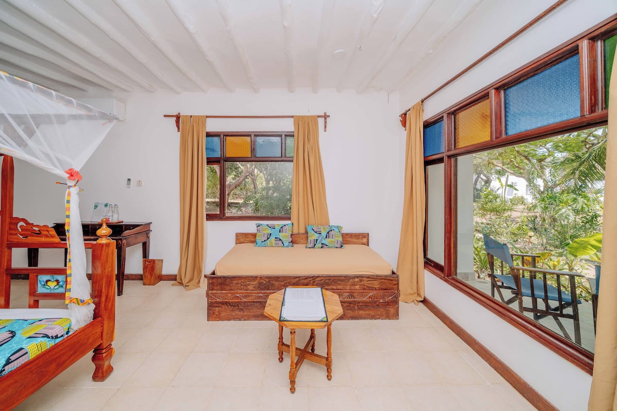 A bedroom with a wooden bed, a small wooden table, a window with curtains, and a view of greenery outside.