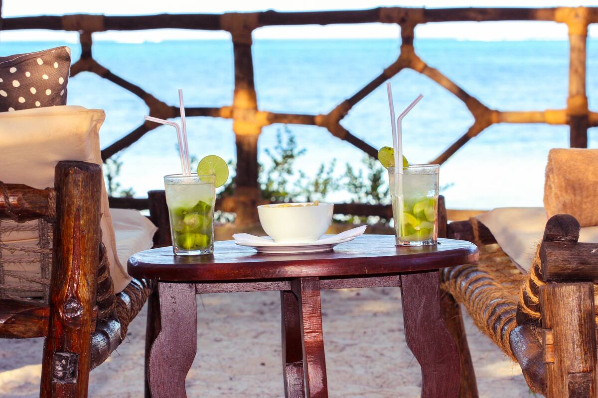 A wooden table with a bowl and two glasses of a green beverage, set against a view of the sea.