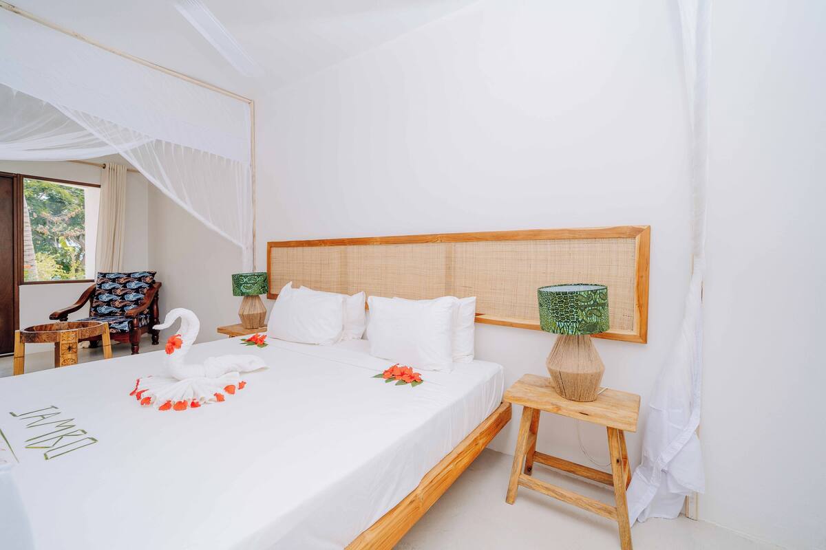 A neatly made bed with white linens, a wooden headboard, and a decorative swan. There are two bedside tables with lamps, a chair, and a window with curtains.