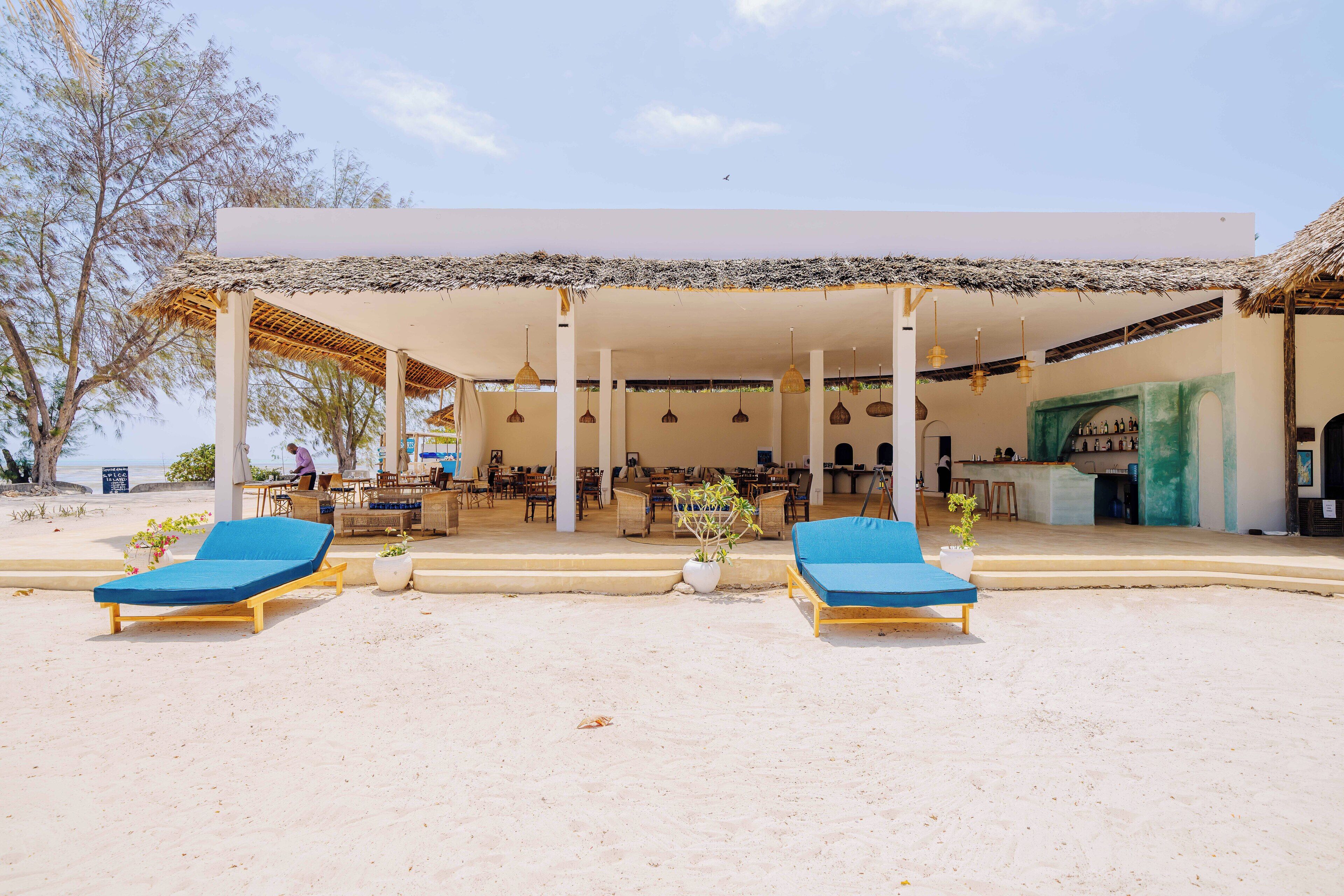 Un hotel frente a la playa con dos sillas azules de descanso, una zona de estar cubierta y un bar o sala de estar.