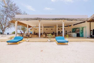 Un hotel frente a la playa con dos sillas azules de descanso, una zona de estar cubierta y un bar o sala de estar.