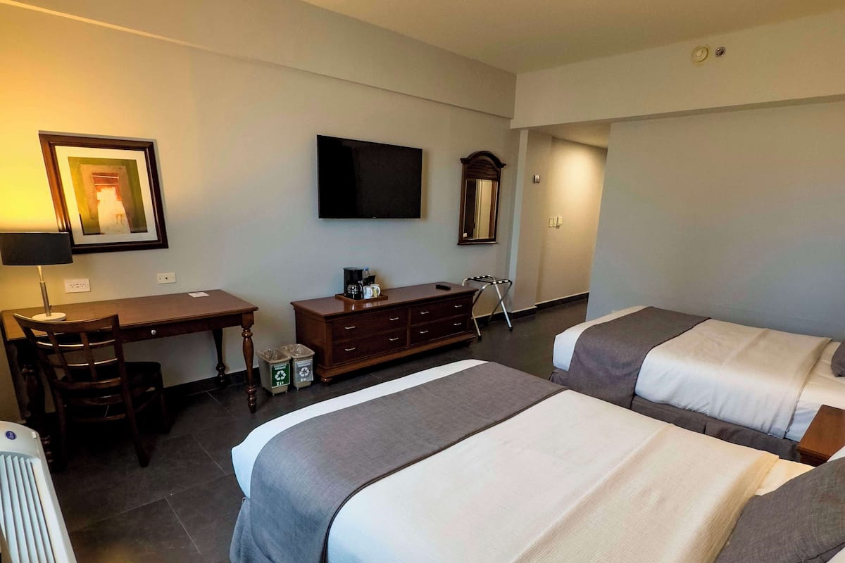 Quarto de hotel com duas camas, uma escrivaninha, uma cadeira, cômoda, televisão e espelho.