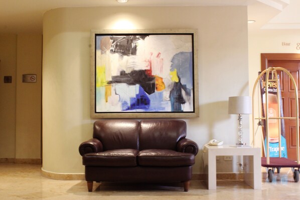 A lobby with a brown leather sofa, a framed abstract painting, a small table with a lamp, and a luggage cart. - Best Western Plus Los Mochis (Los Mochis)