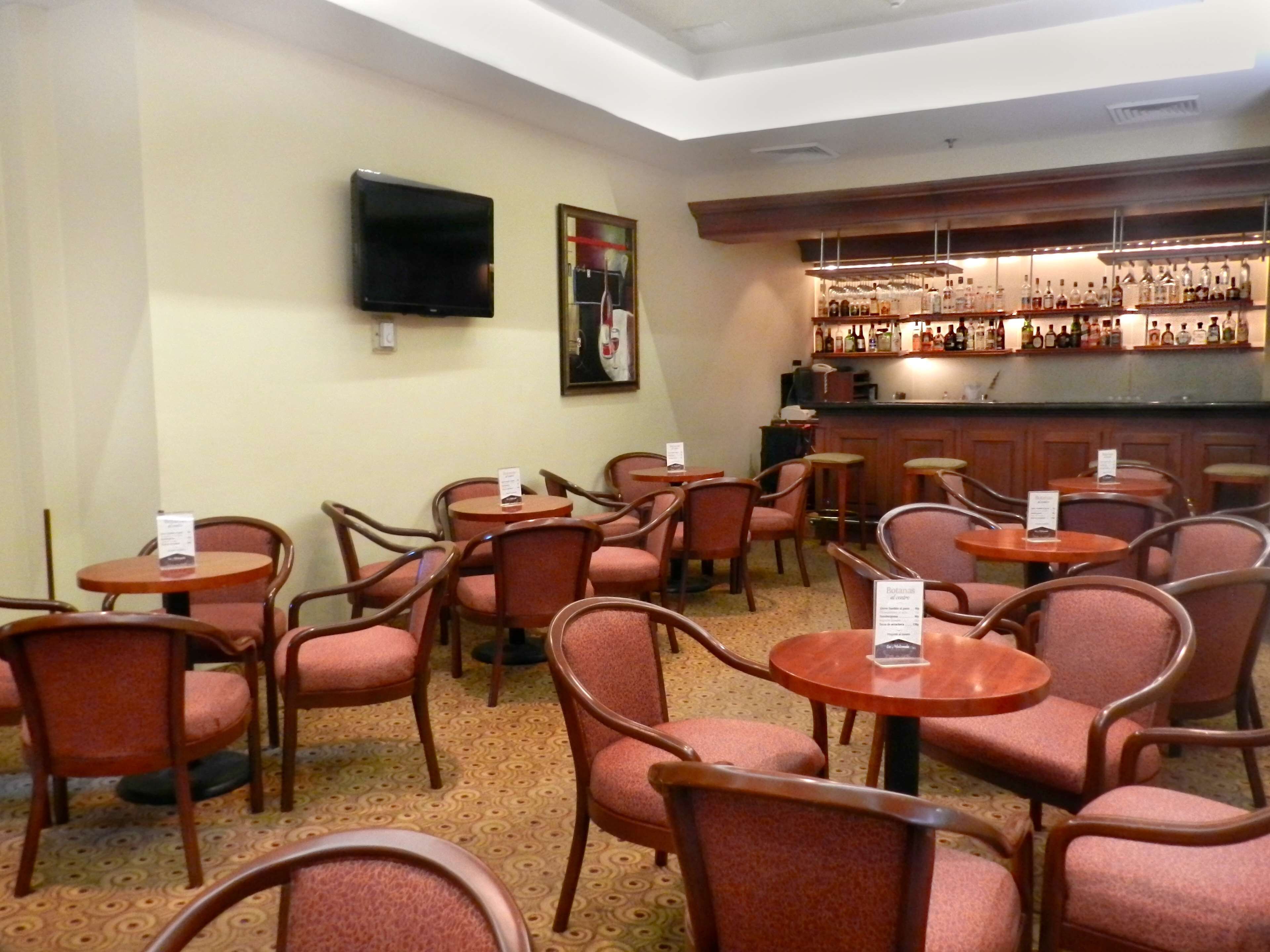 A hotel lobby with multiple round tables and chairs, a bar area with various liquor bottles, a flat-screen TV mounted on the wall, and a framed artwork above the bar.