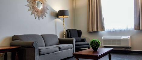 A living room with a grey sofa, a black armchair, a wooden coffee table, a floor lamp, a mirror, and a window with curtains.