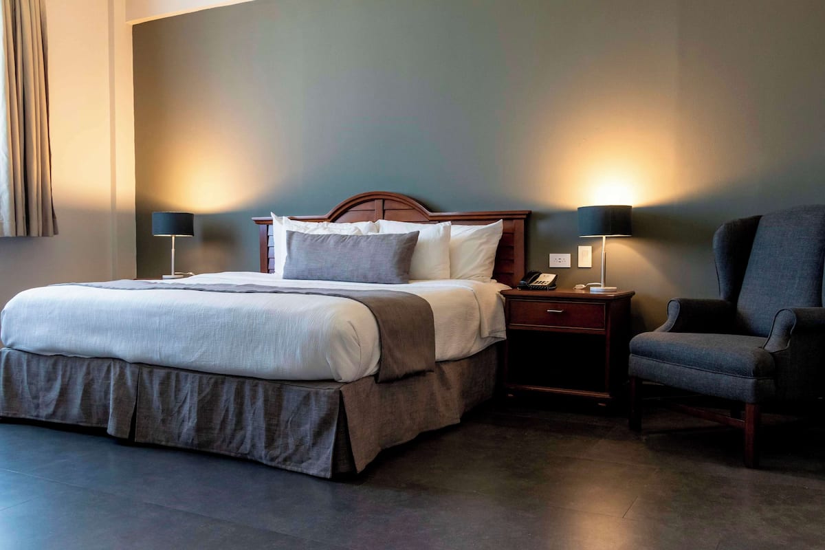 Quarto de hotel com uma cama grande, duas mesas de cabeceira com luminárias, uma poltrona e um criado-mudo com telefone.