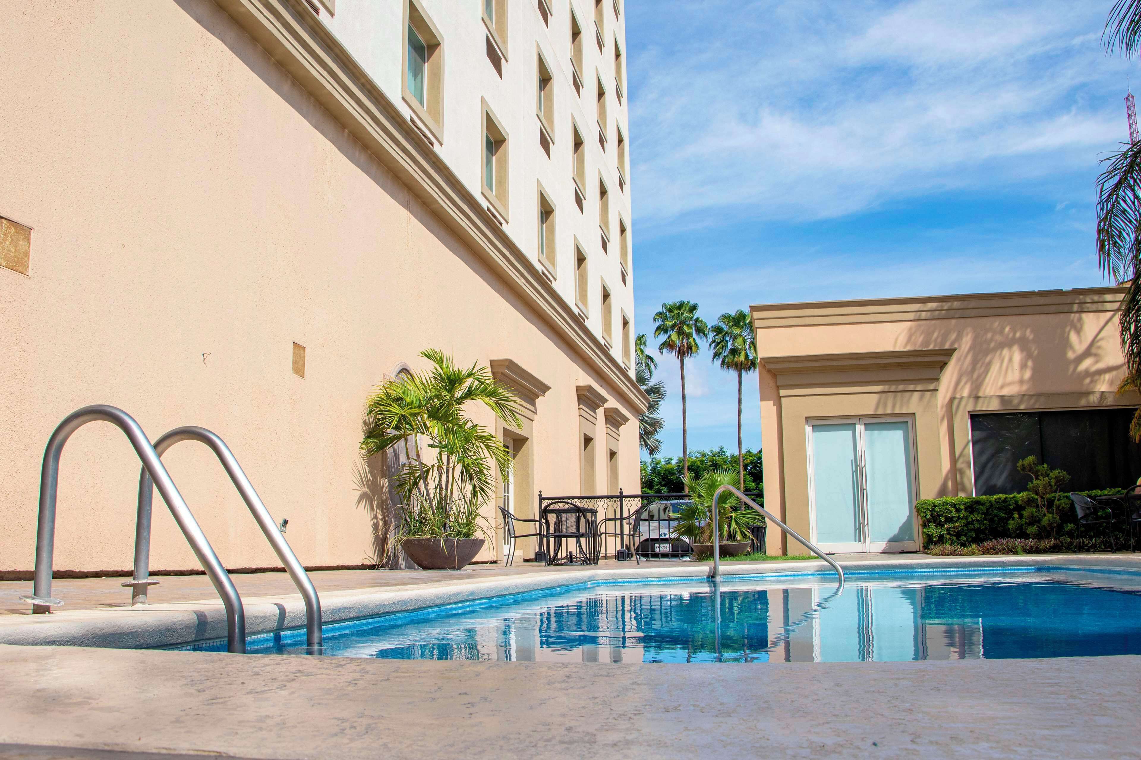 A swimming pool with metal ladders, a palm tree, and a building with a beige facade.