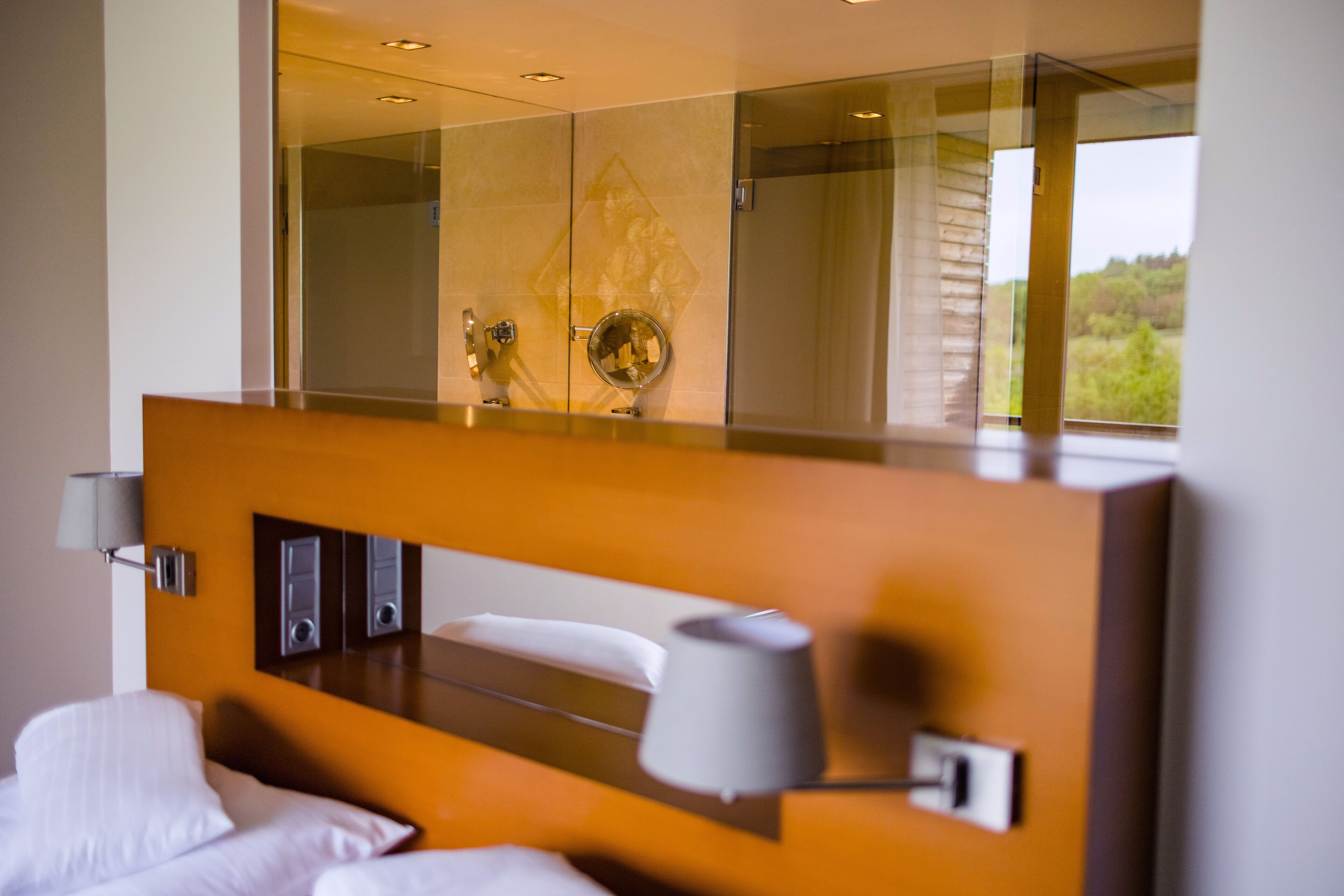 A hotel room with a large mirror, a bed with white linens, a bedside table with a lamp, and a view of greenery outside.