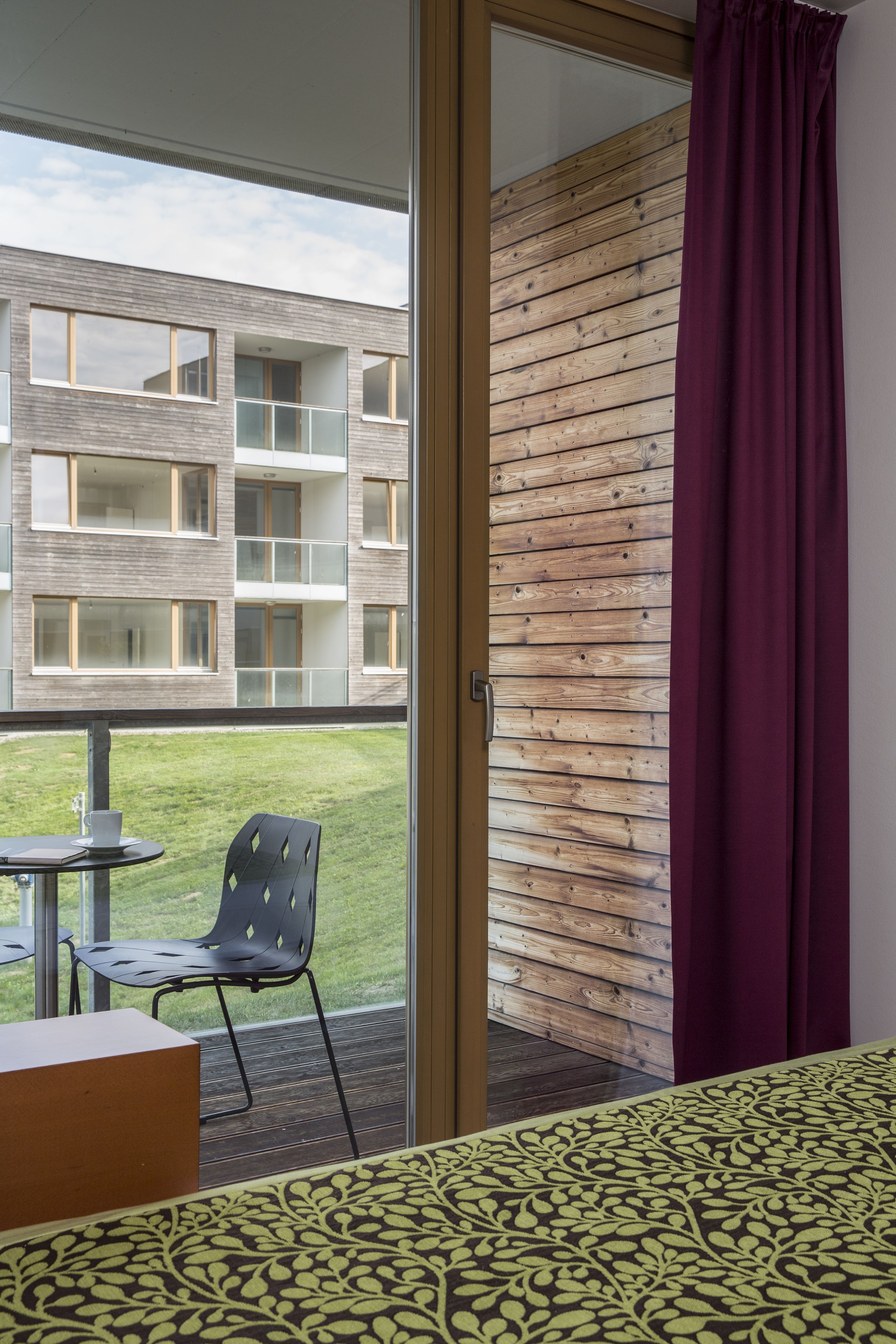 A hotel room with a balcony, a wooden wall, and a view of a building with multiple windows.