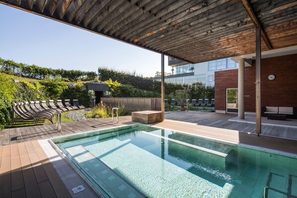Un área de piscina moderna al aire libre con una terraza de madera, sillas de descanso y un espacio de estar cubierto.