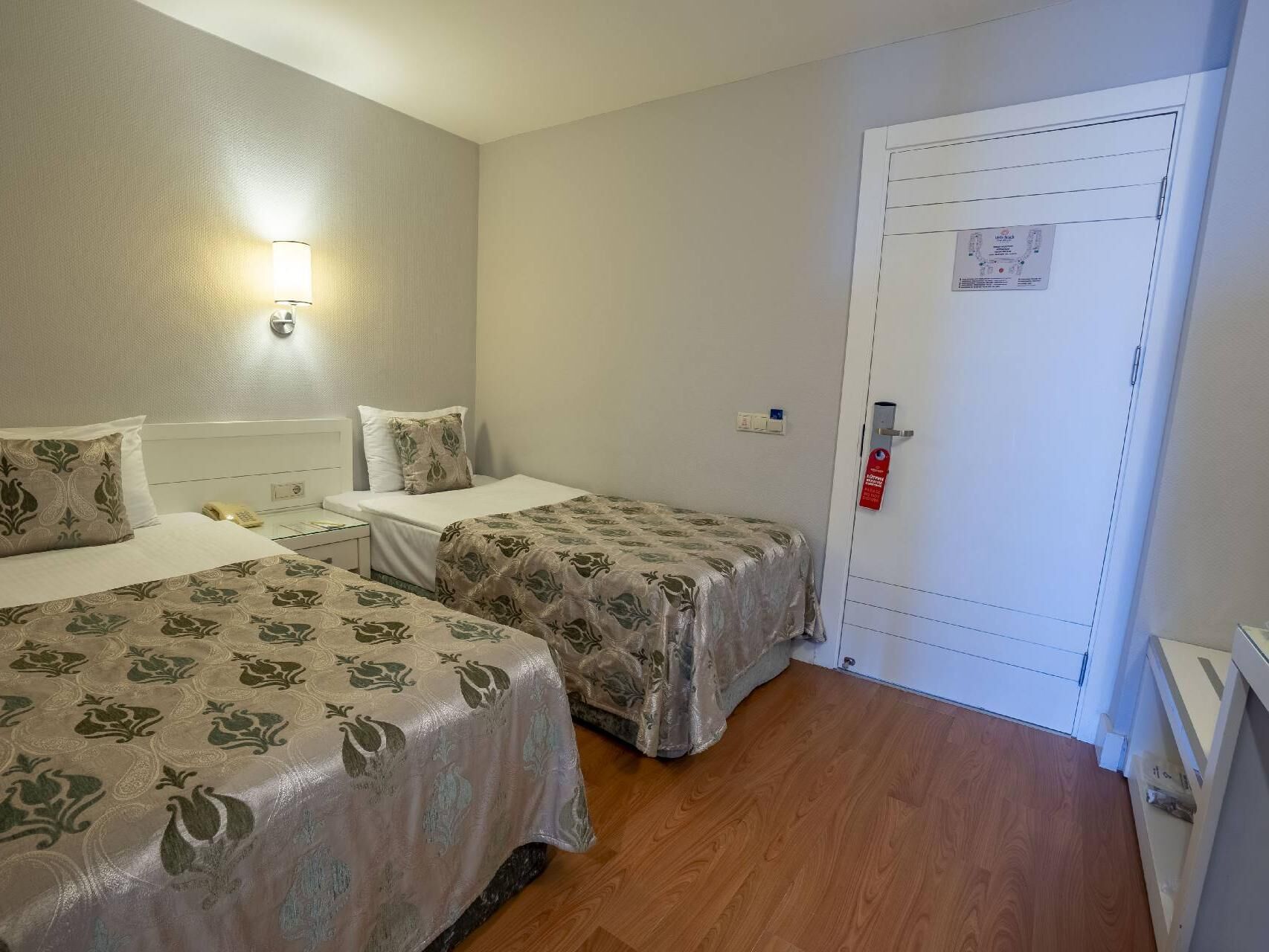 Habitación de hotel con dos camas, una mesita de noche, una lámpara, una puerta con detector de incendios y suelo de madera.