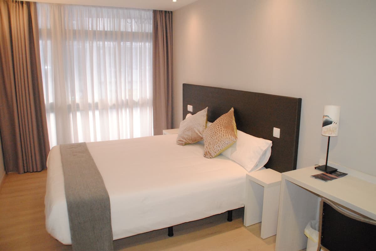 A neatly made bed with a grey blanket, two pillows, a bedside table with a lamp, and a window with curtains.
