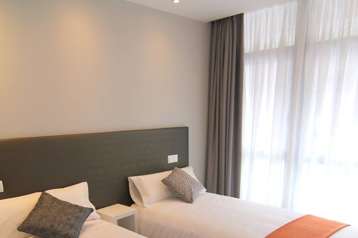 Two single beds with white linens and orange throw blankets, a dark headboard, and a window with sheer curtains.