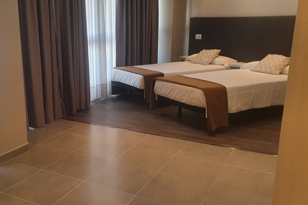 A hotel room with two beds, a chair, and a view of the outdoors through curtains.