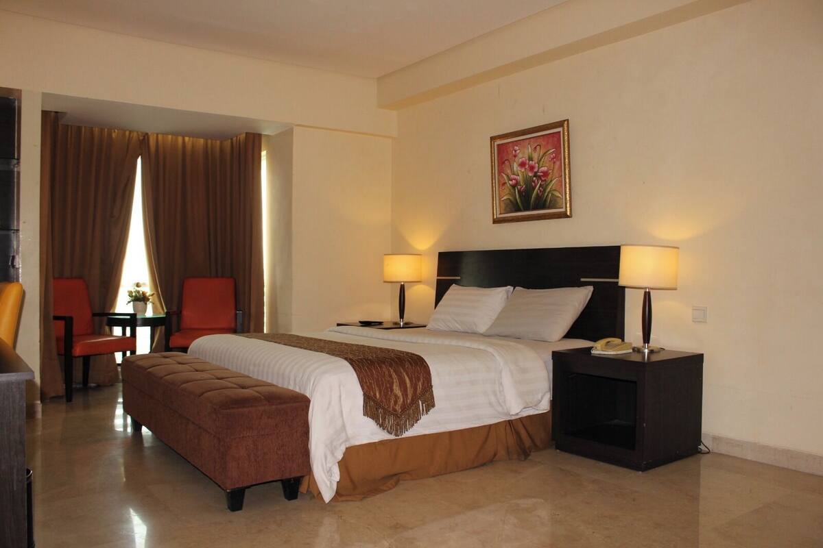 A hotel room with a bed, two bedside tables with lamps, a chair, a small table with a vase, and a painting on the wall.