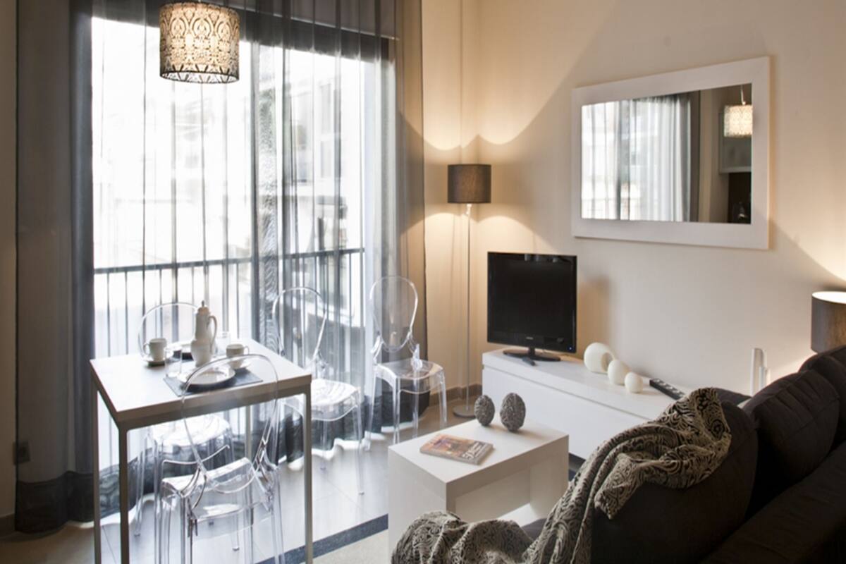 A modern living room with a dining area, a flat-screen TV, and a large window with curtains.