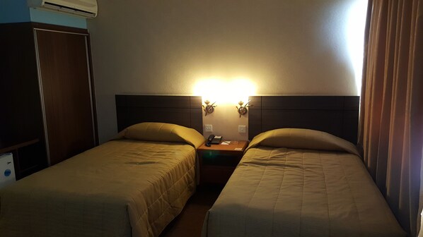 Una habitación de hotel con dos camas individuales, una mesita de noche con lámpara y un armario.