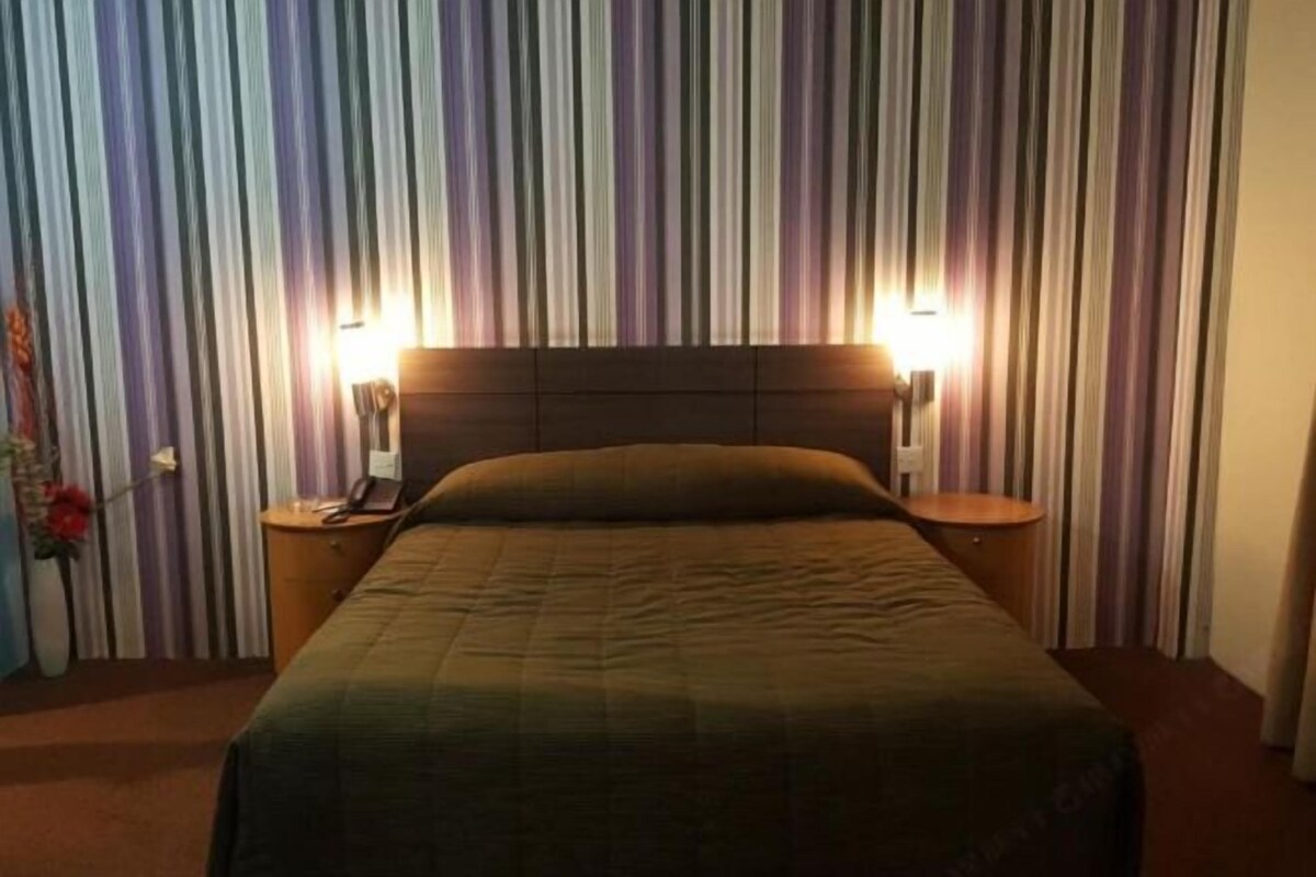 A hotel room with a striped curtain backdrop, a bed with a green blanket, two bedside tables with lamps, and a vase with flowers on one table.