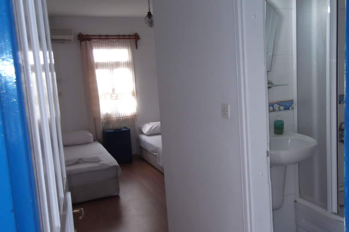 A room with a bed, a small table, a window with curtains, and a bathroom with a sink and mirror.