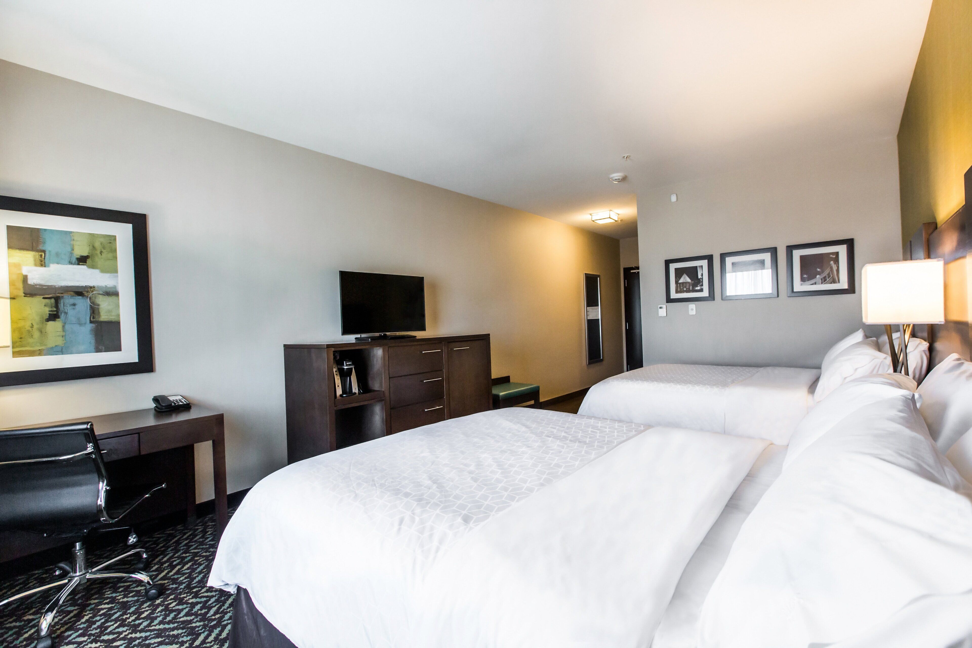 A hotel room with two beds, a desk, a chair, a television, and framed pictures on the wall.