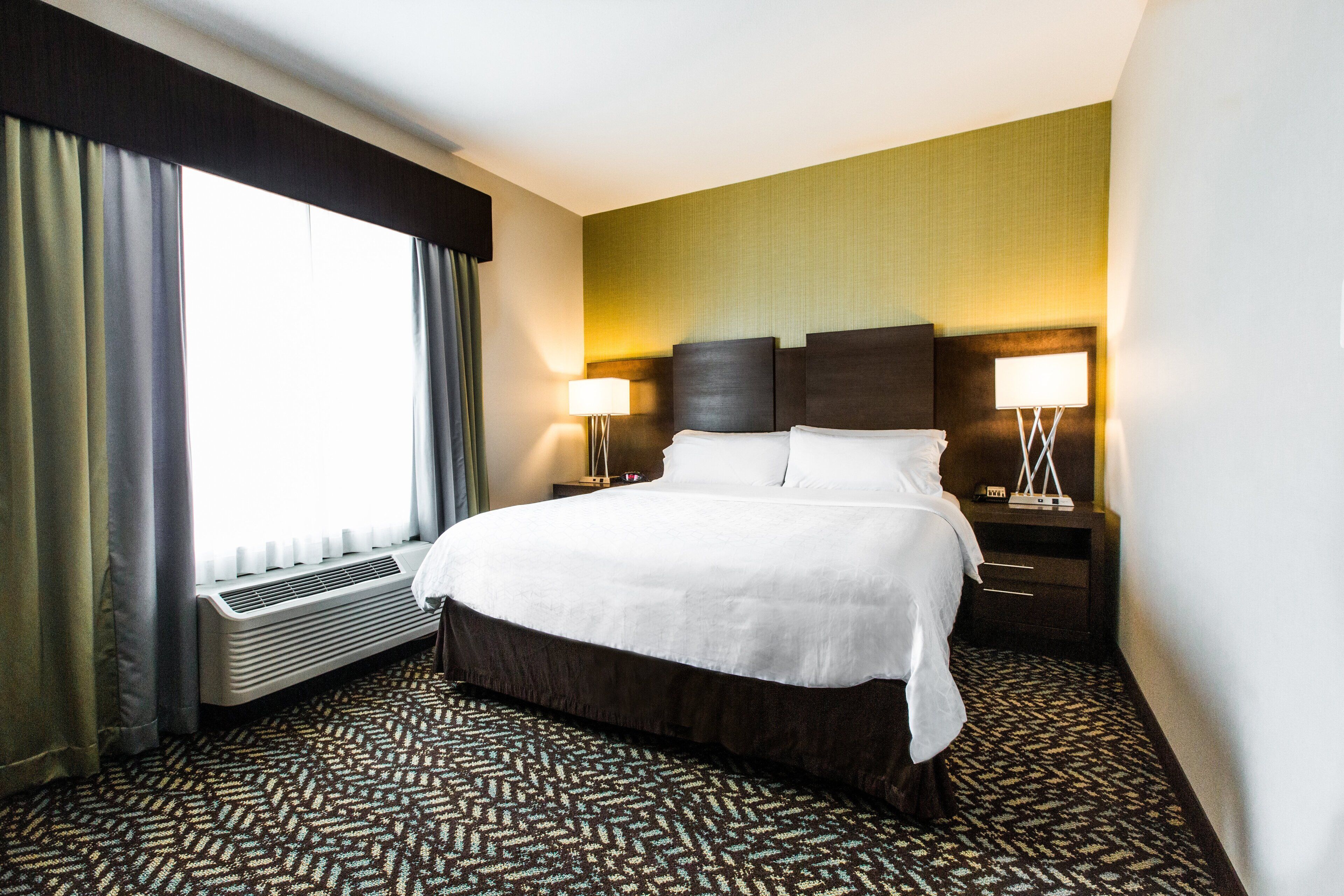 A hotel room with a large bed, two bedside tables with lamps, a window with curtains, and a patterned carpet.